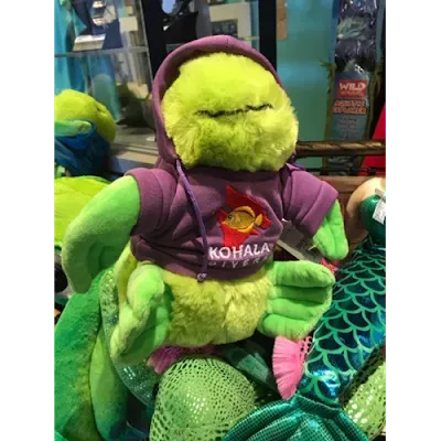 A stuffed frog wearing a purple hoodie is sitting on a pile of stuffed animals.