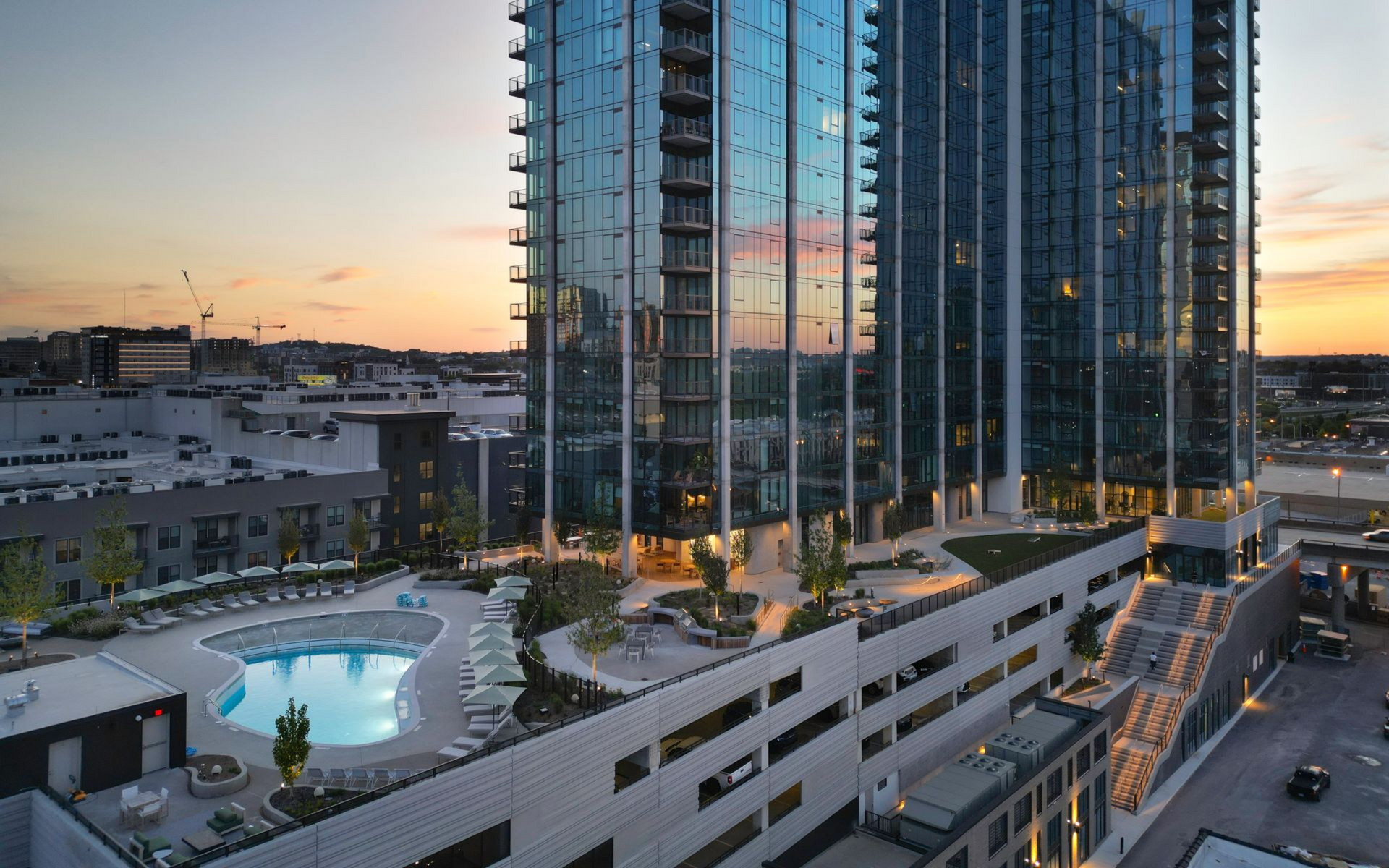 High-rise apartments beside a lit pool at dusk, with city streets and sunset sky in the background
