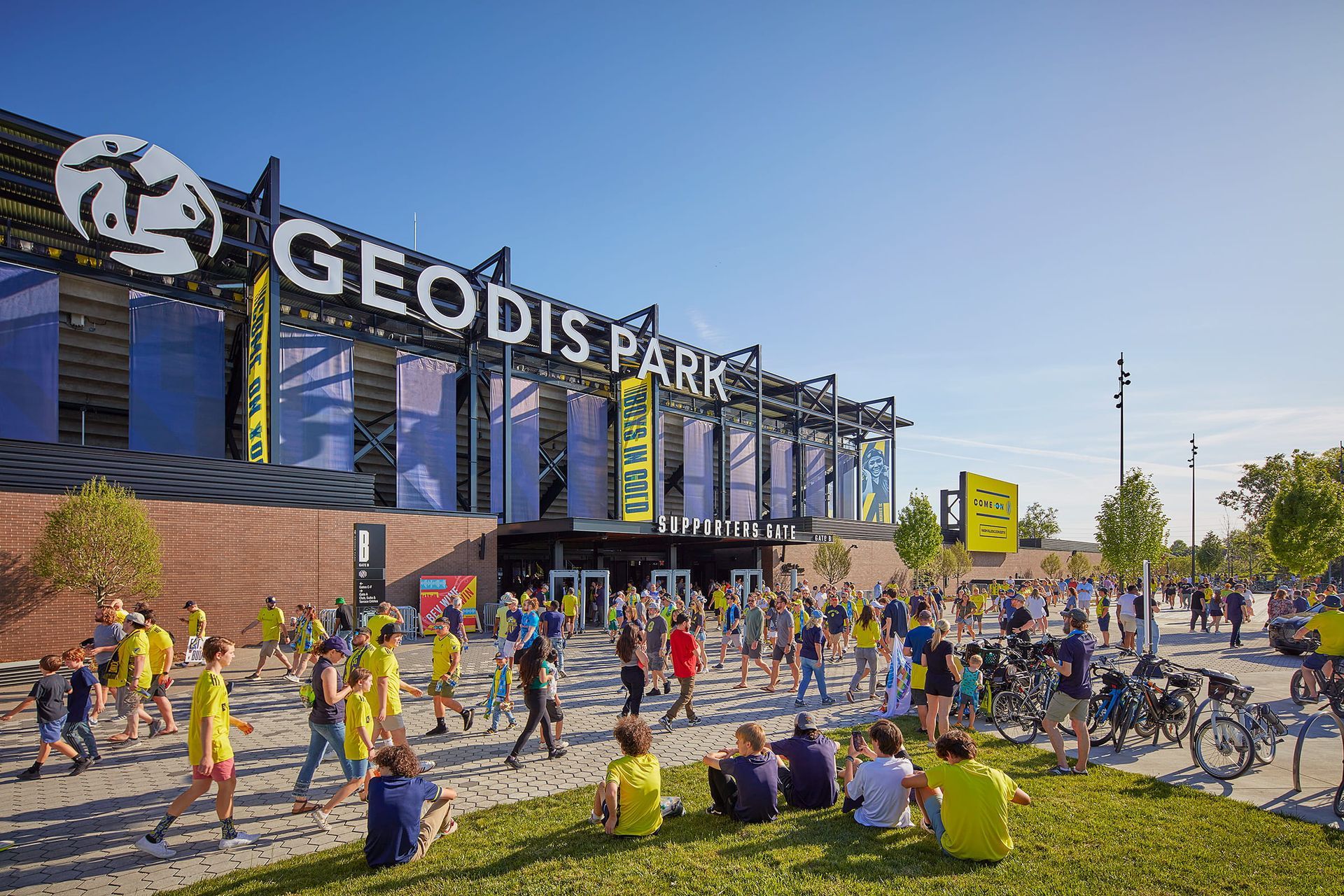 Crowd gathered outside GEODIS Park stadium on a sunny day, with people sitting on the grass and bicycles nearby