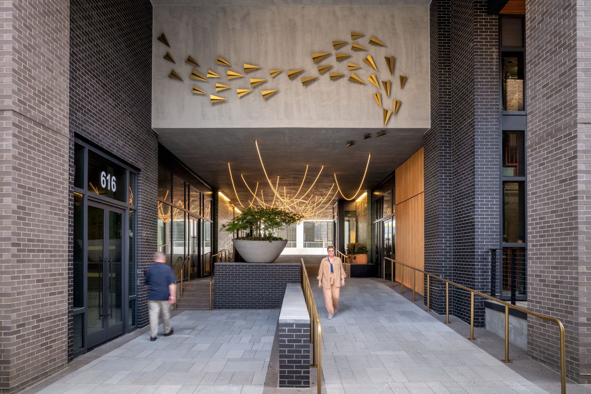 Modern building entrance with brick walls, recessed lighting, and people walking through a covered walkway