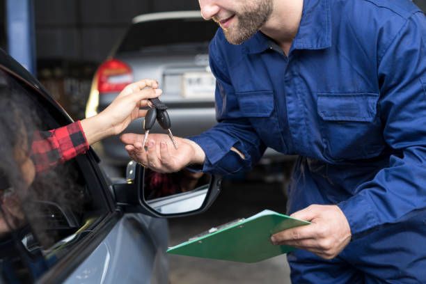 A mechanic in blue coveralls receives car keys from a person in a vehicle at a shop.