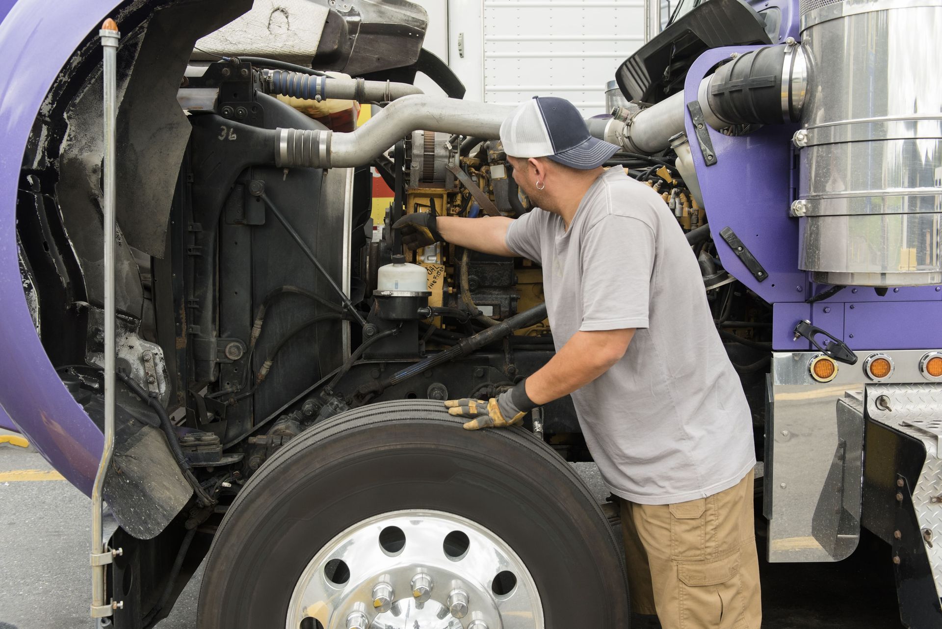 Mechanic inspecting engine of a purple semi-truck; wearing a cap, gloves, and khaki pants.