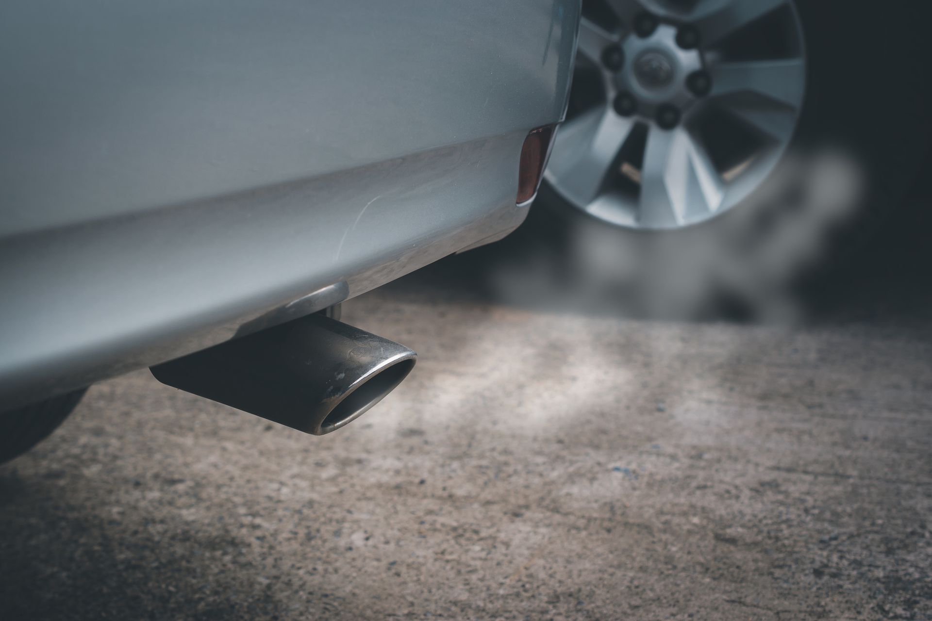 Exhaust pipe of a silver car emitting smoke on a concrete surface, with a blurred tire in the background.
