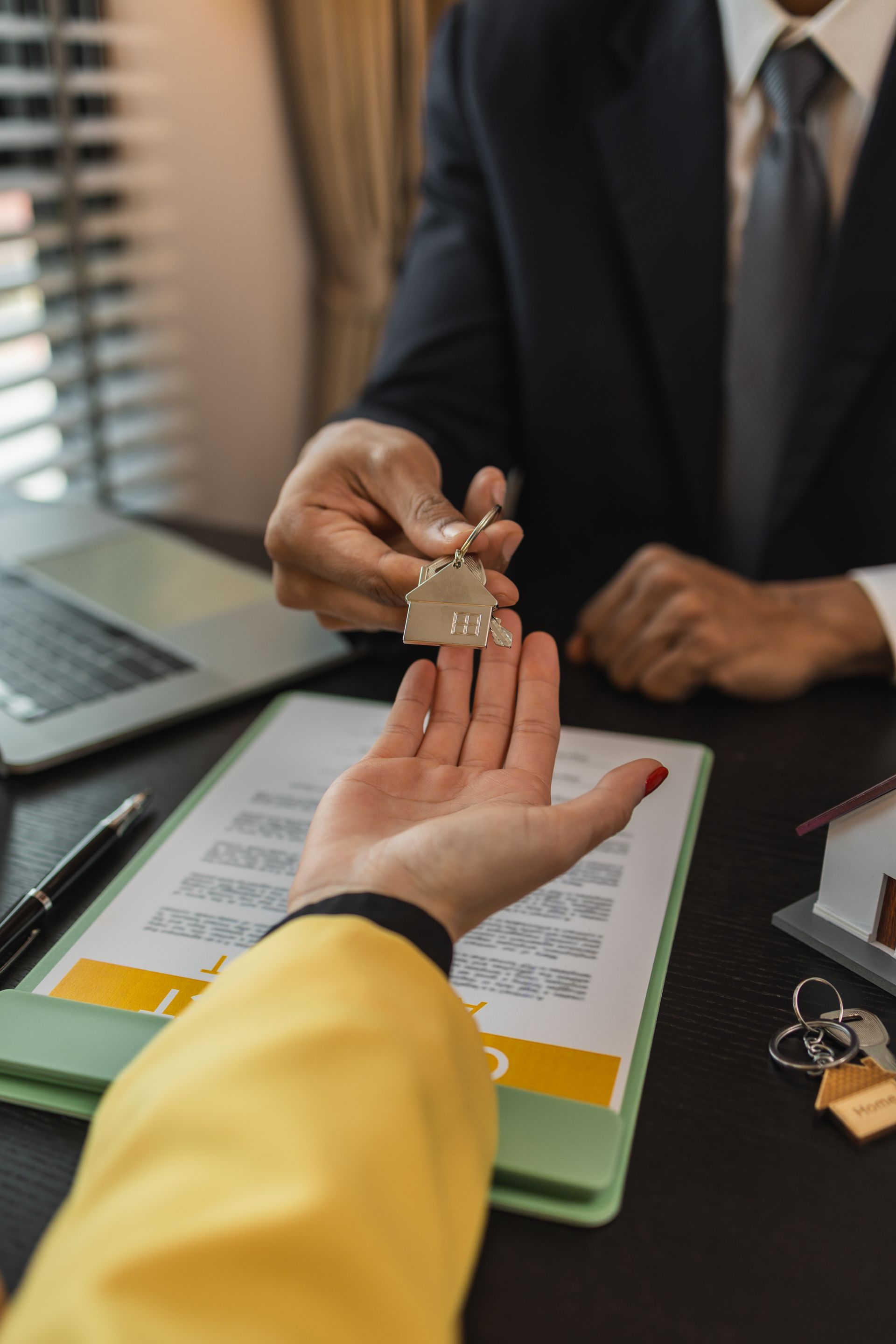 A person in a suit hands house-shaped keys to another person over a desk with a contract and a miniature model home. A person in a suit hands house-shaped keys to another person over a desk with a contract and a miniature model home.