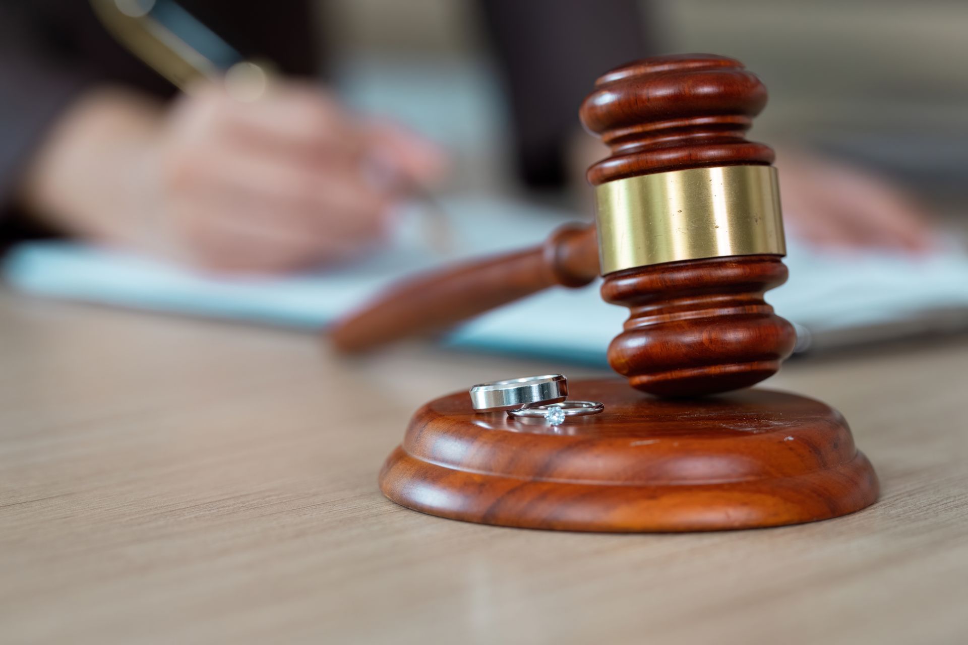 A wooden judge's gavel rests on its base alongside two wedding rings, with a person writing in the background.