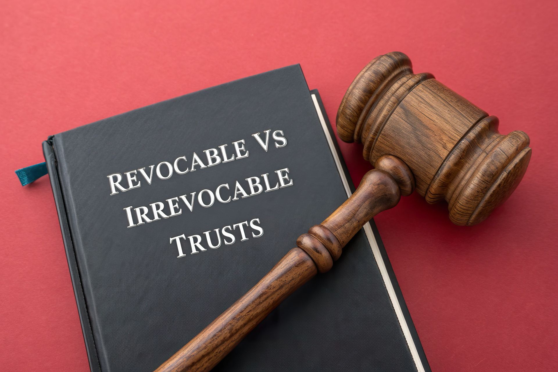 A wooden gavel lies on top of a black book titled Revocable Vs Irrevocable Trusts, set against a red background.