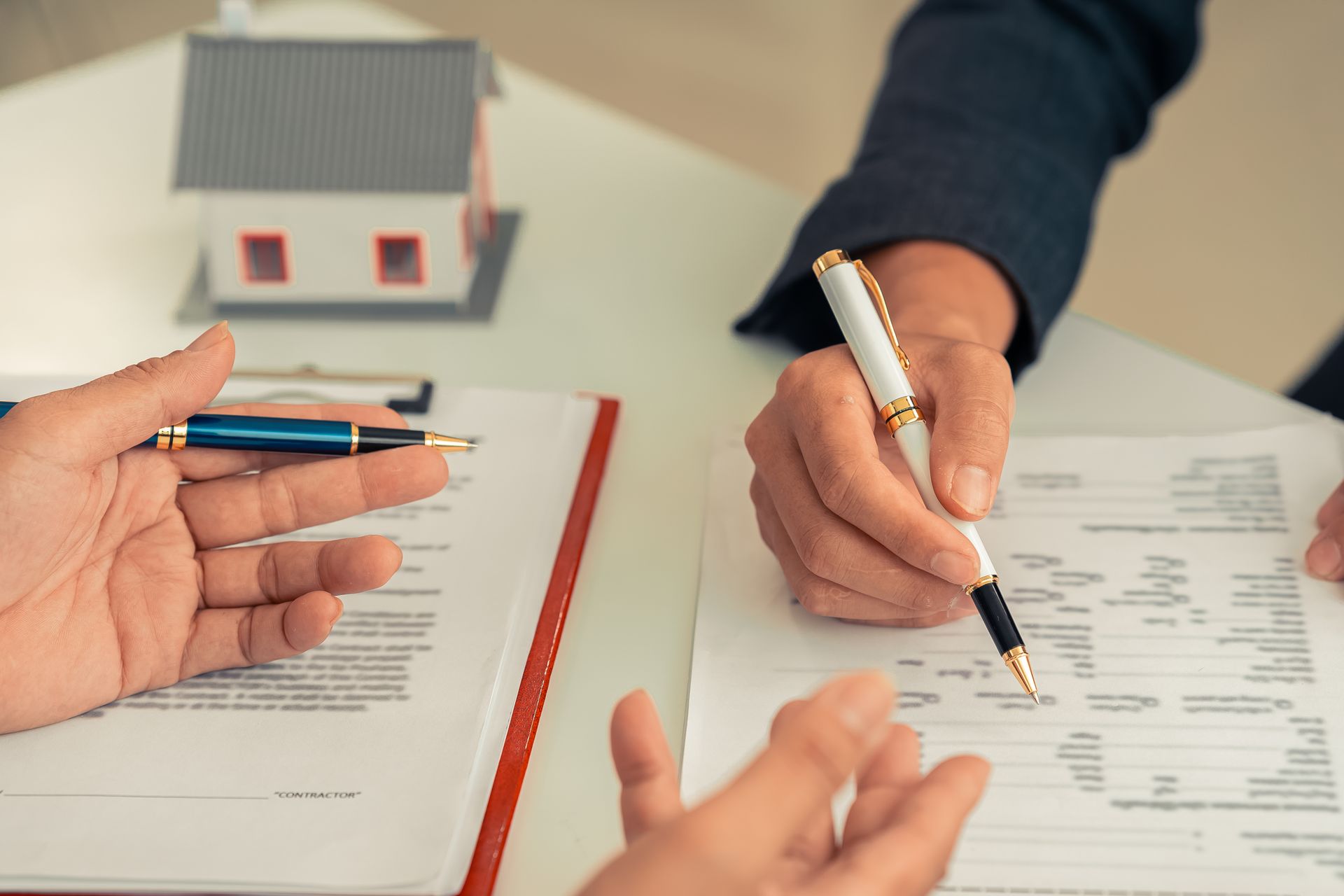 Close-up of two people signing real estate paperwork next to a miniature house model on a white table. Close-up of two people signing real estate paperwork next to a miniature house model on a white table.
