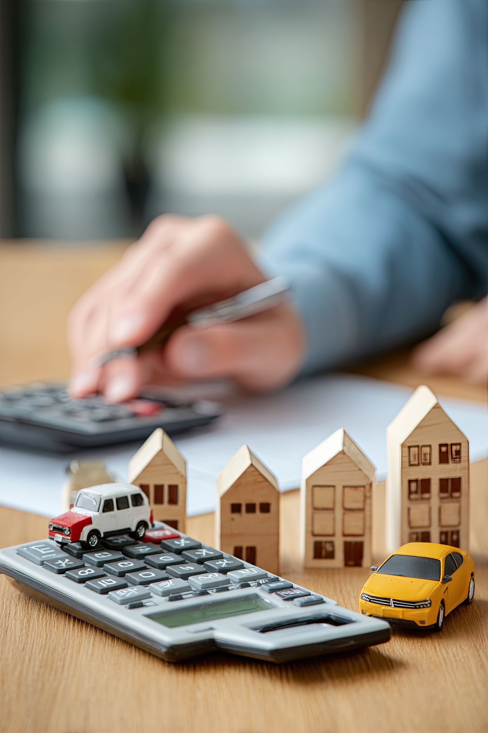 A person uses a calculator and pen next to miniature wooden house models and toy cars on a wooden table.