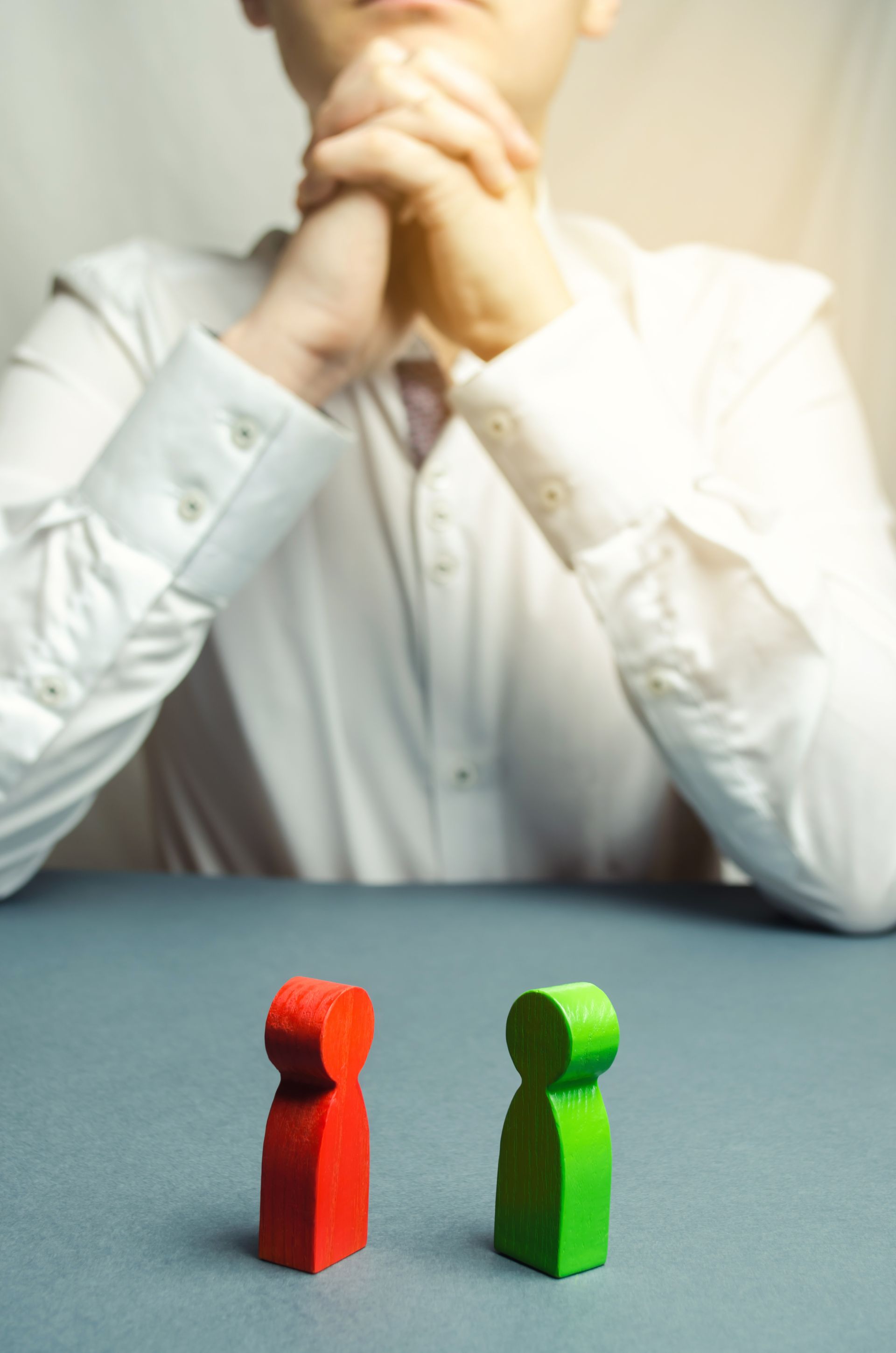 A person sits with hands clasped behind a red and a green wooden figure placed on a table.