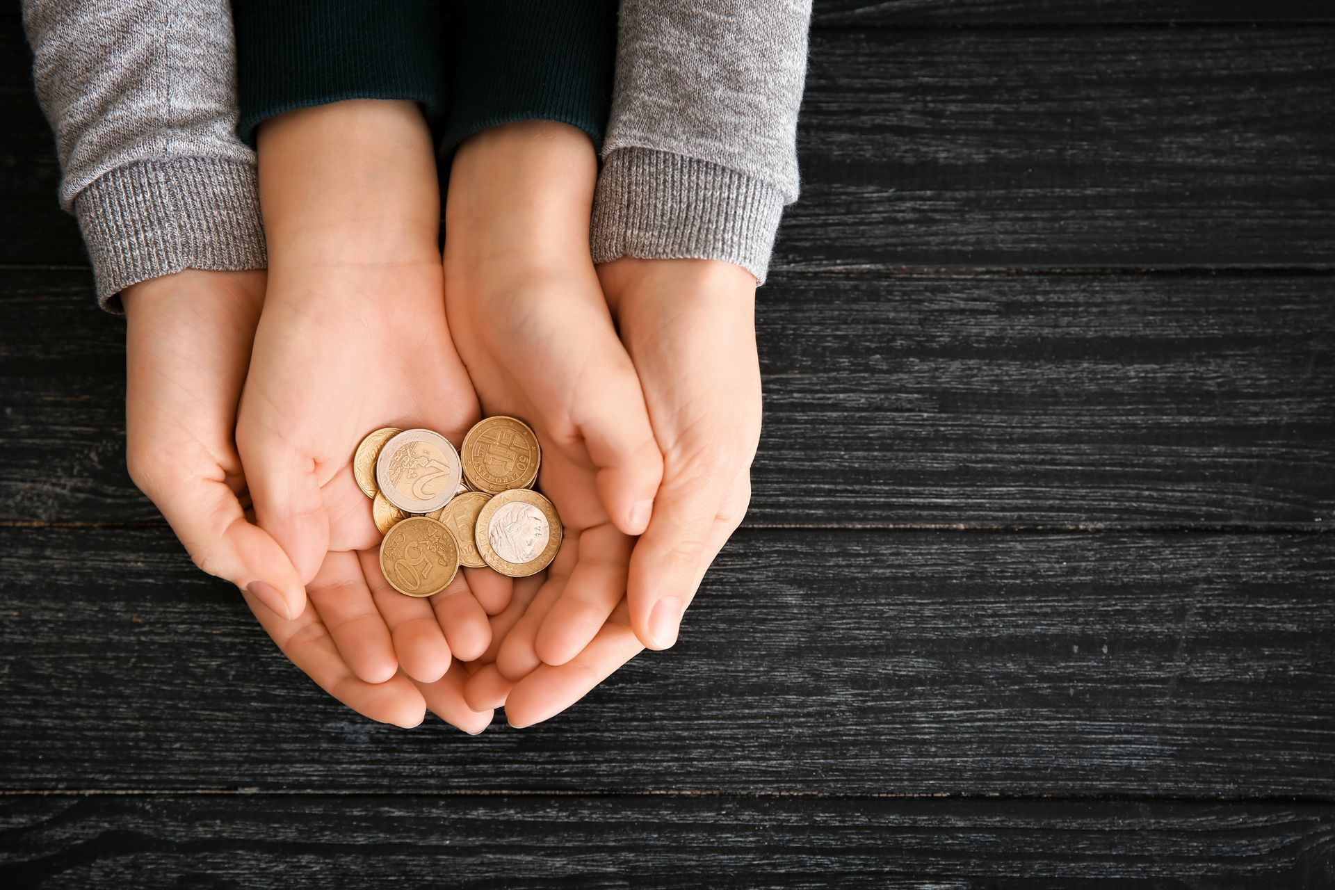 Two pairs of hands held together against a dark wooden background, cradling a small pile of gold-colored coins.