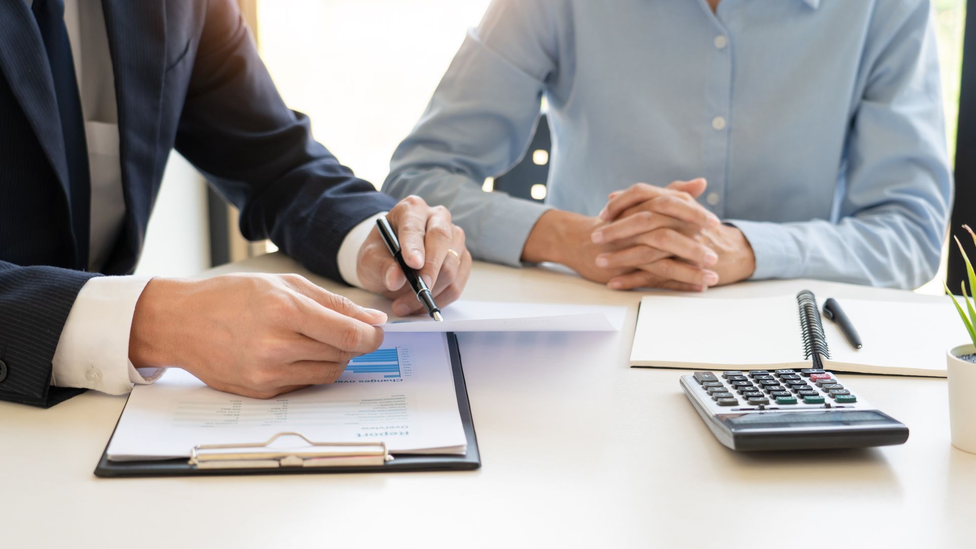 Two people in professional attire sit at a desk reviewing documents and working with a calculator.