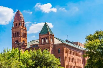 A historic red brick building with a tall, domed tower and green shingled roofs under a bright blue sky with clouds.