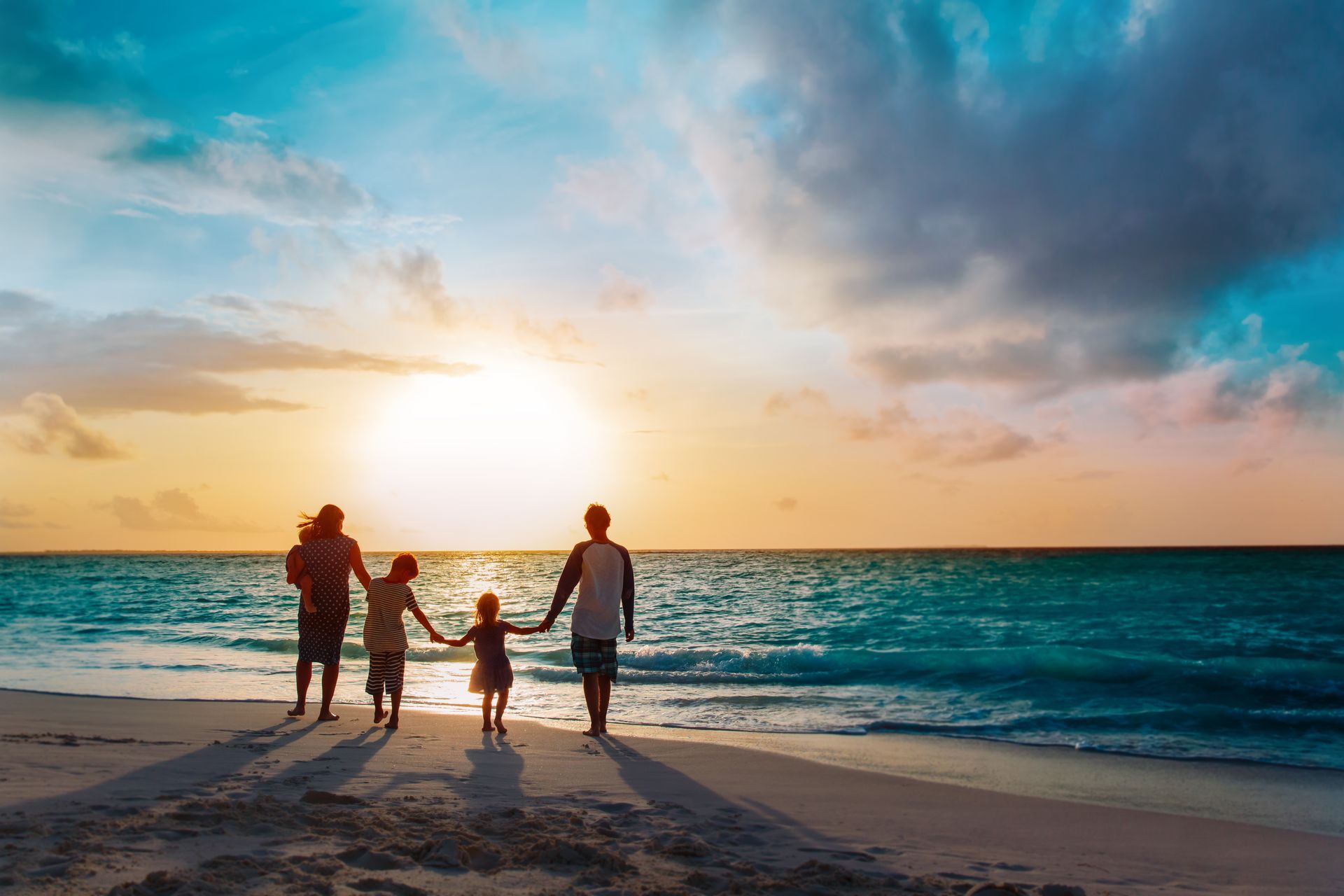 A family holds hands while walking along a sandy beach during a vibrant, sunlit sunset over the ocean.