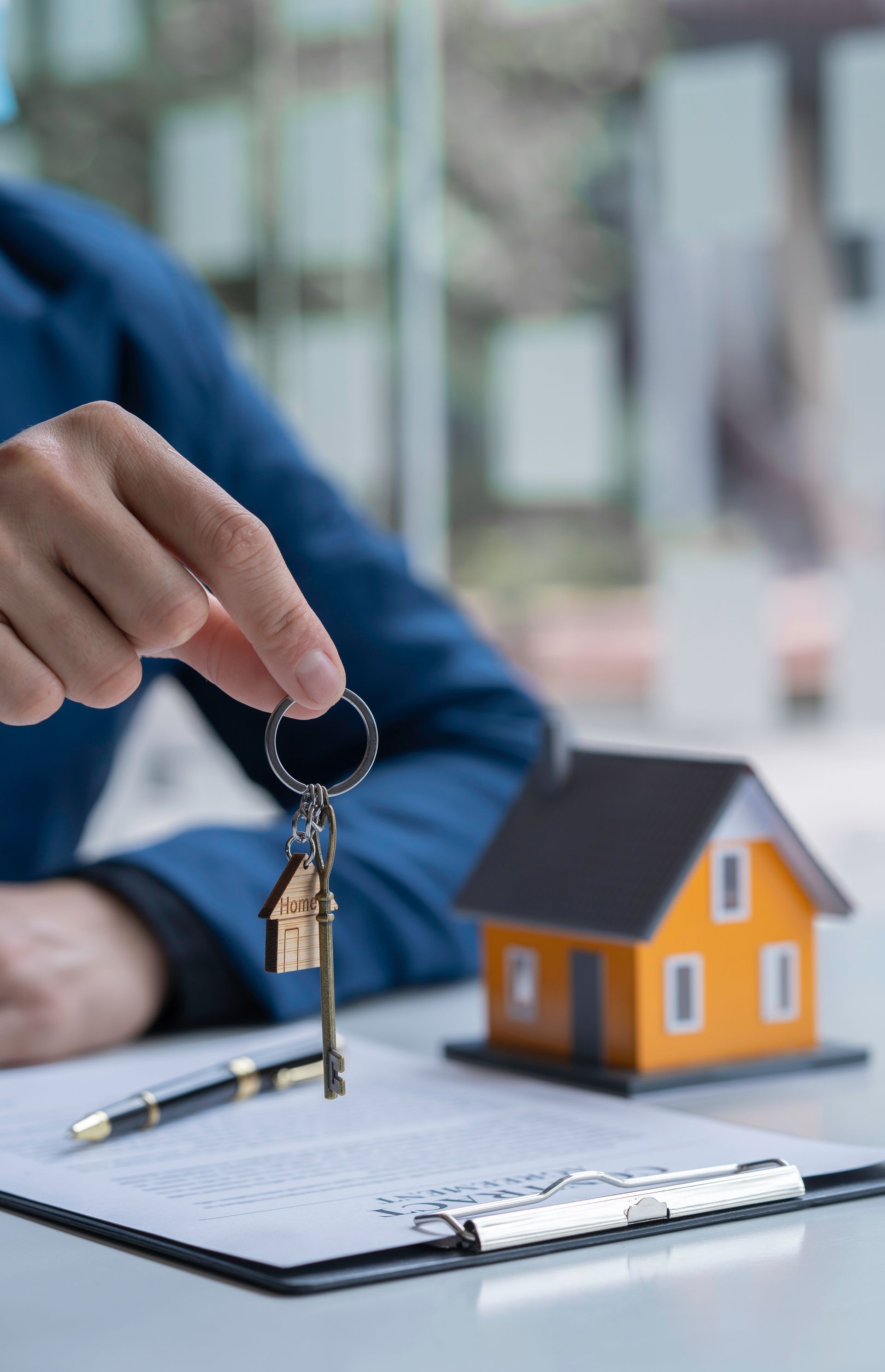 A person in a blue jacket holds house-shaped keys over a document, next to a small orange house model on a desk. A person in a blue jacket holds house-shaped keys over a document, next to a small orange house model on a desk.