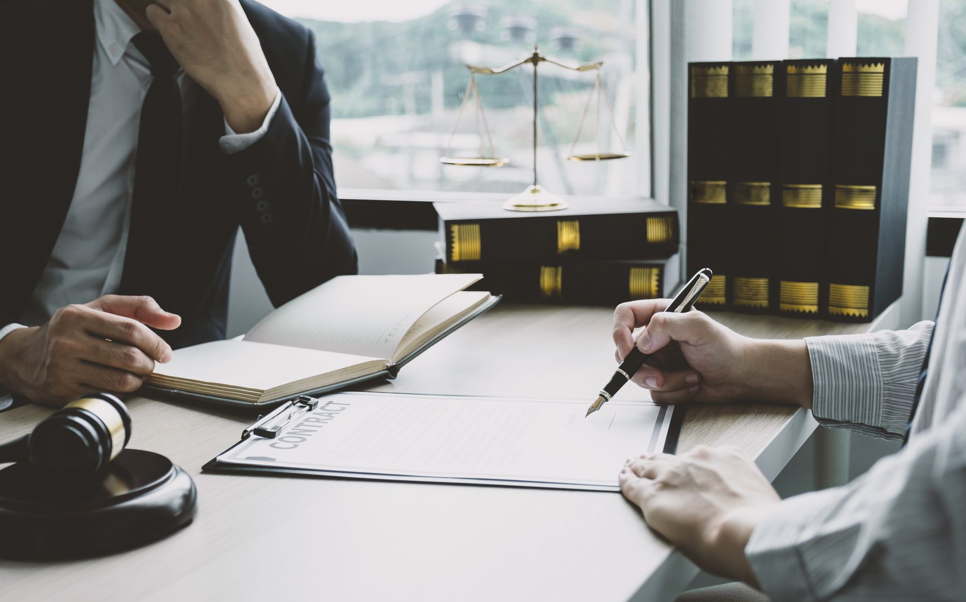 A lawyer and client sit at a desk with law books, a gavel, and a balance scale while the client signs a document.