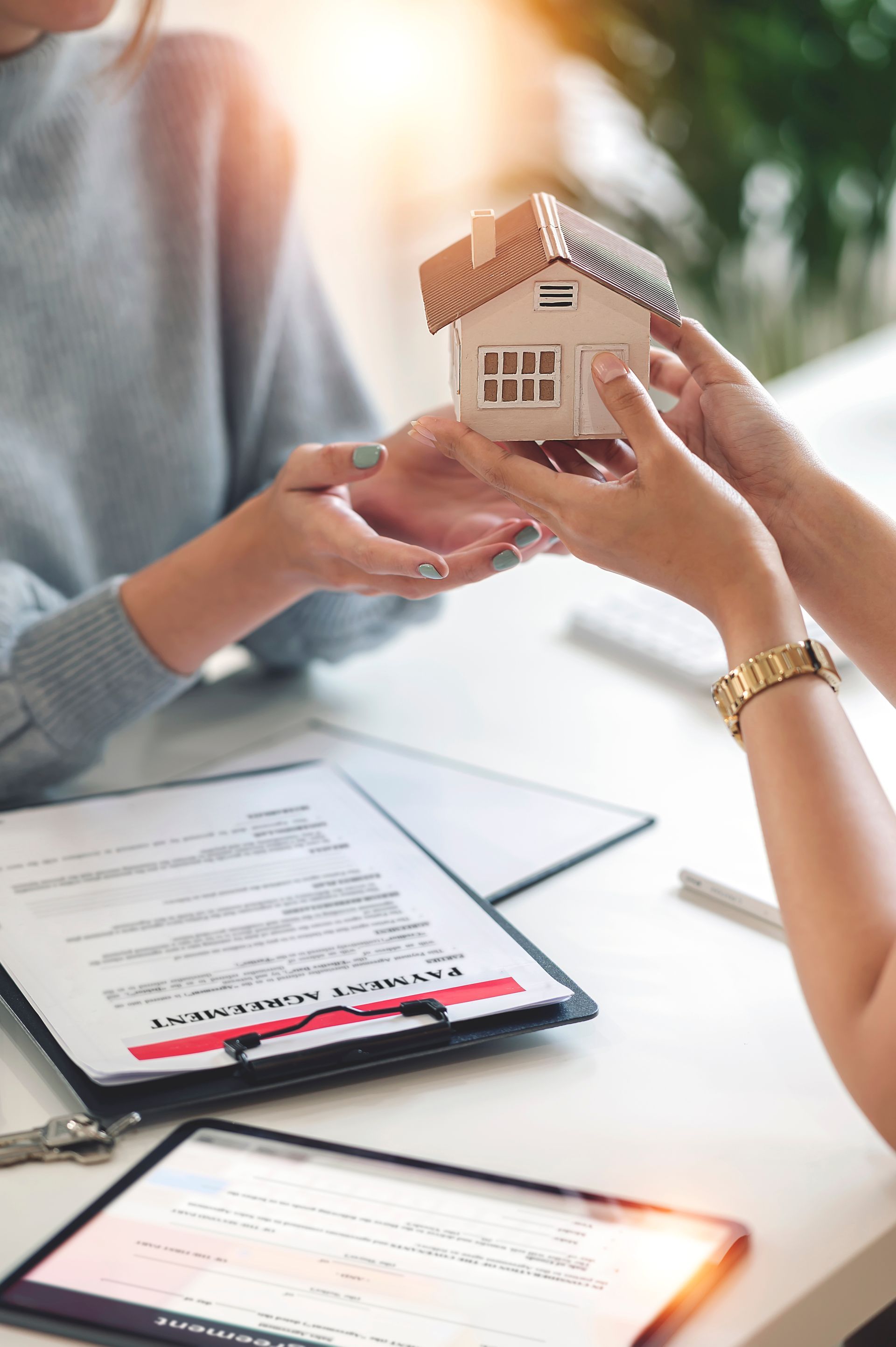 Two people exchange a small model house over a table with documents and a tablet, signifying a real estate transaction. Two people exchange a small model house over a table with documents and a tablet, signifying a real estate transaction.