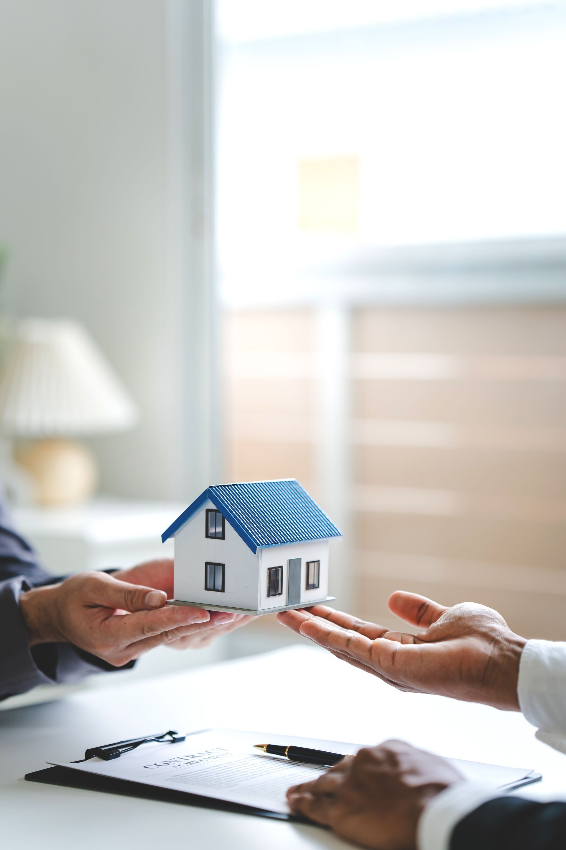 Two people exchange a small model house over a desk with paperwork, representing a real estate or insurance transaction. Two people exchange a small model house over a desk with paperwork, representing a real estate or insurance transaction.