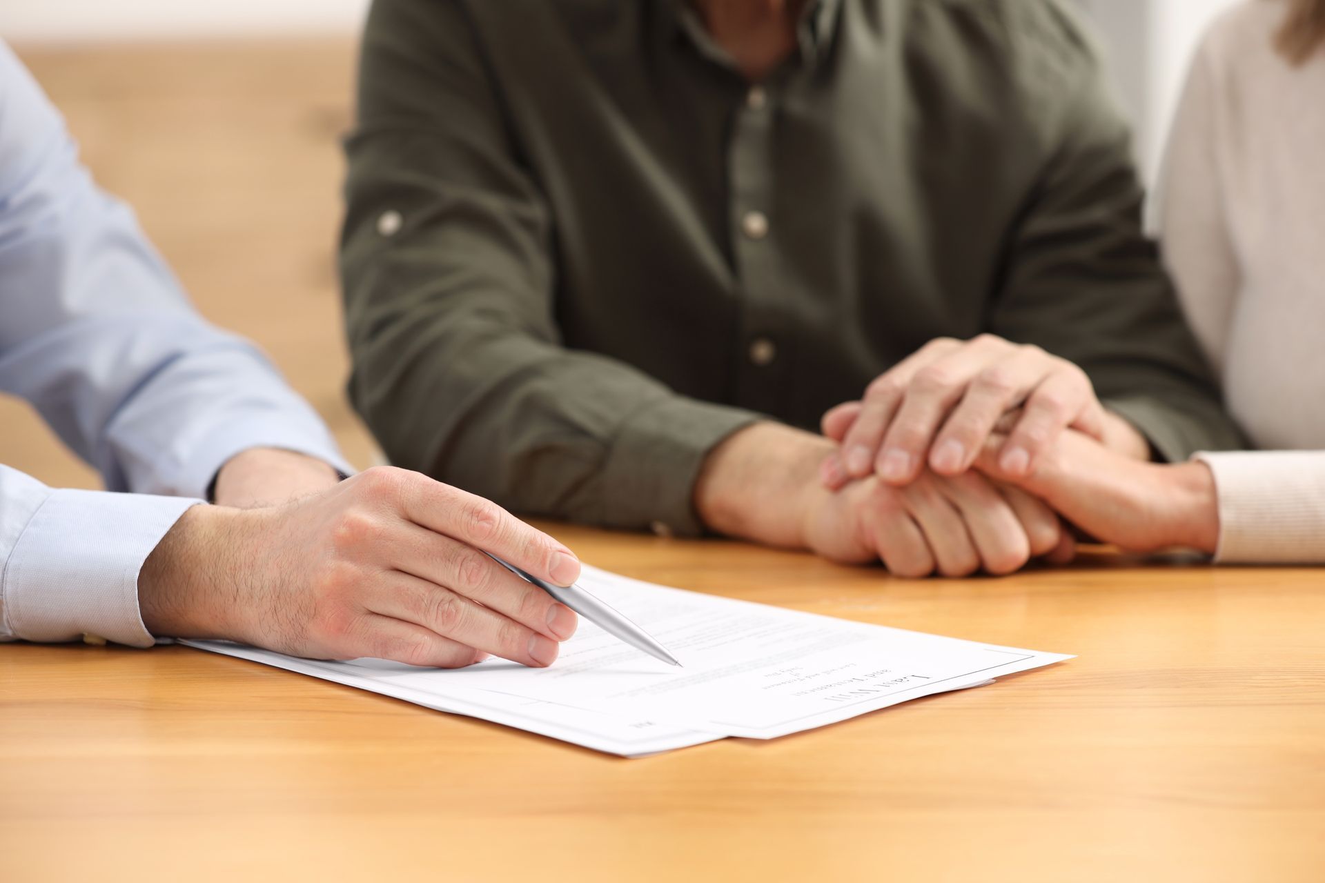 A professional points to a document on a wooden table, while two people hold hands in the background. A professional points to a document on a wooden table, while two people hold hands in the background.