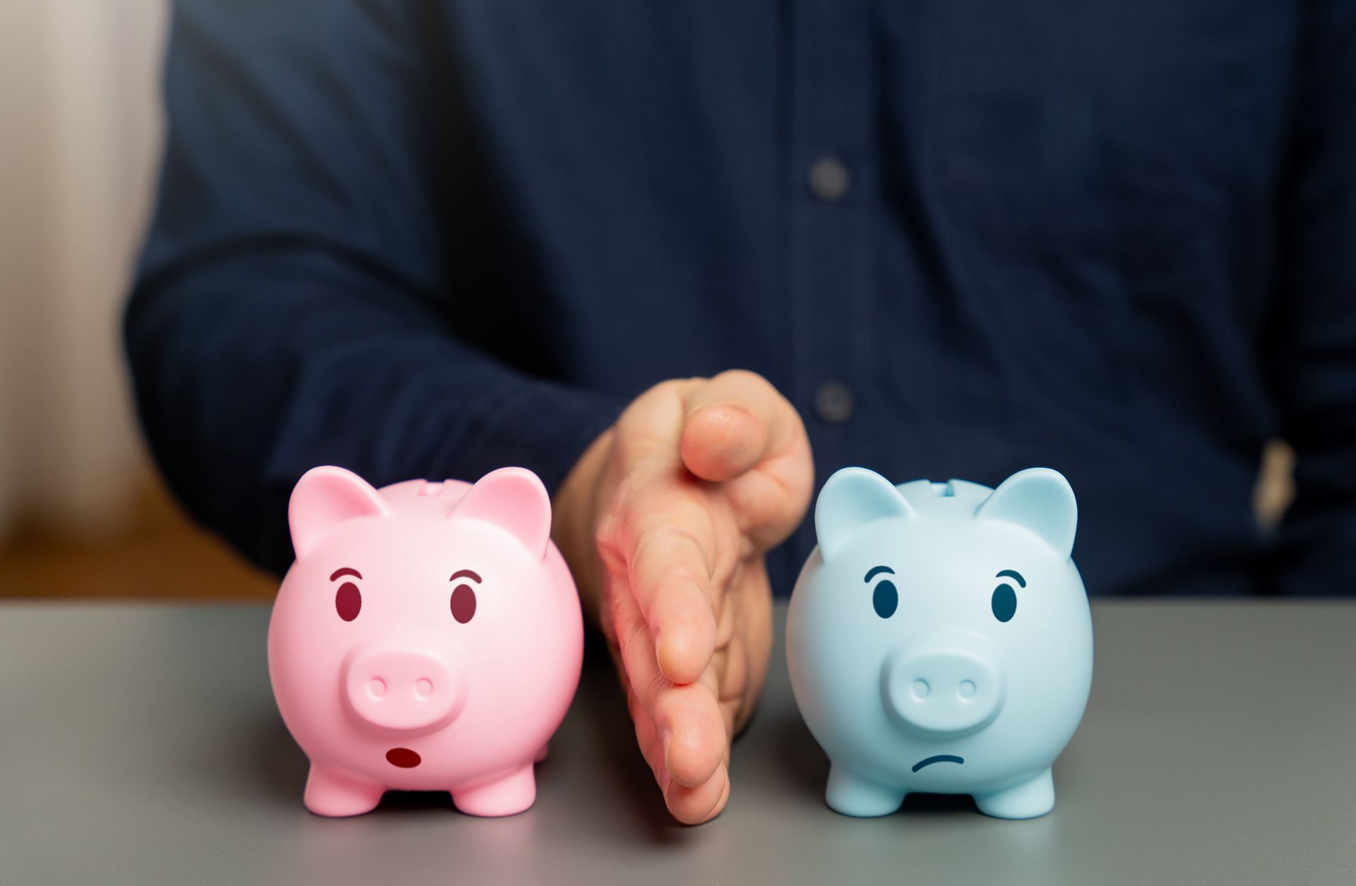 A person separates a pink piggy bank from a blue piggy bank with a sad face, using their hand as a partition.