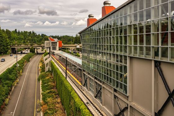 A modern transit station with a large glass facade sits beside a highway and a green, tree-lined landscape under a sky.
