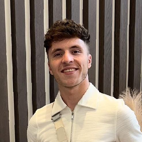 A young man in a white shirt is smiling in front of a wooden wall.