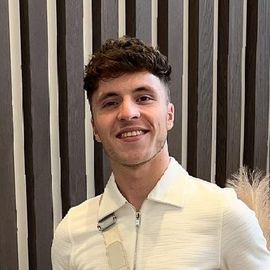 A young man in a white shirt is smiling in front of a wooden wall.