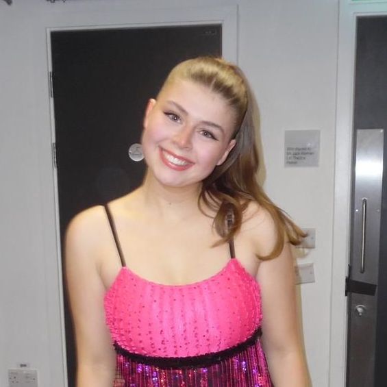 A woman wearing a black tank top and a necklace is smiling in a dark room.