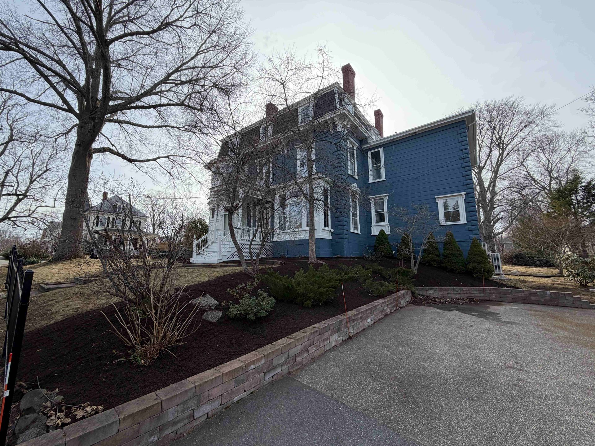Blue house with white trim beside a driveway and leafless trees on a cloudy day