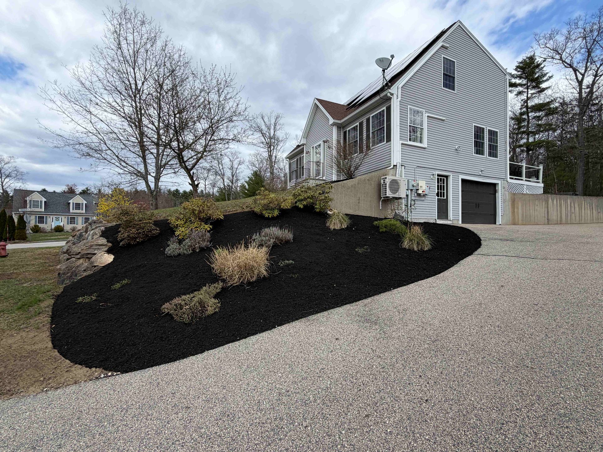 Gray house on a hillside with black mulch landscaping and a gravel driveway under an overcast sky