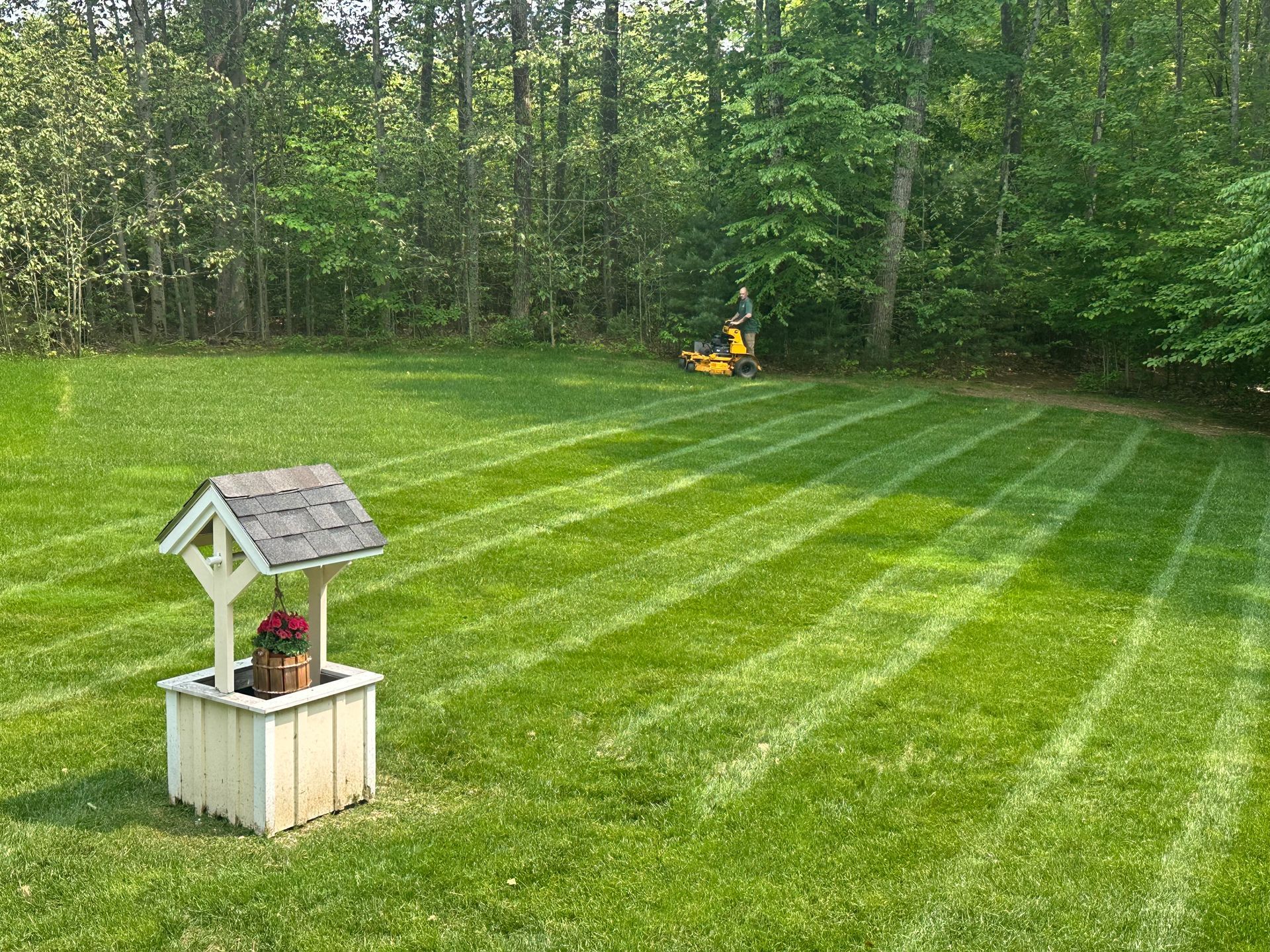 Well-shaped lawn with mowing lines and a decorative well. A yellow lawn mower is in the background near trees.