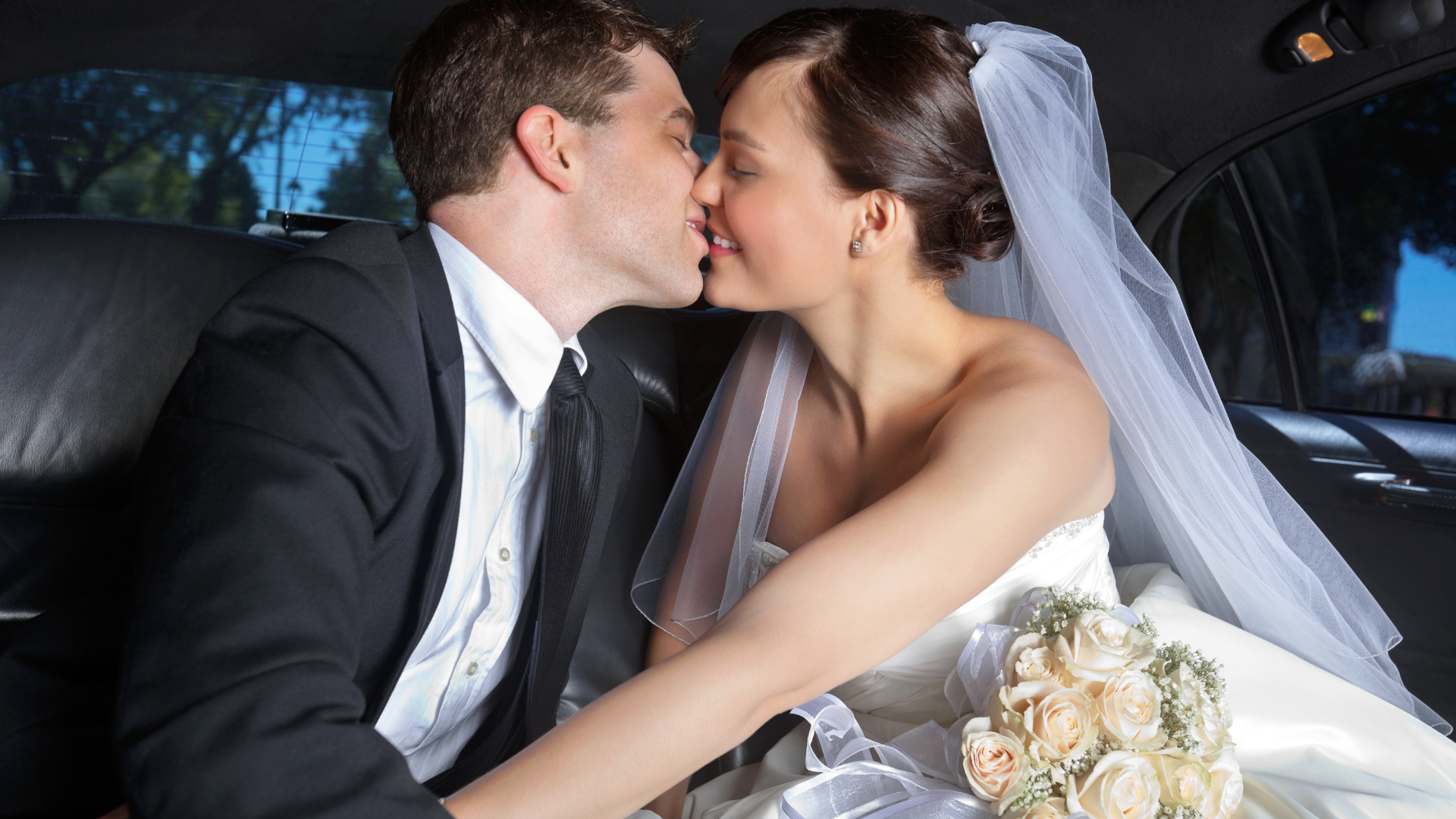 Bride and groom kissing in a car, with a white veil and bouquet visible