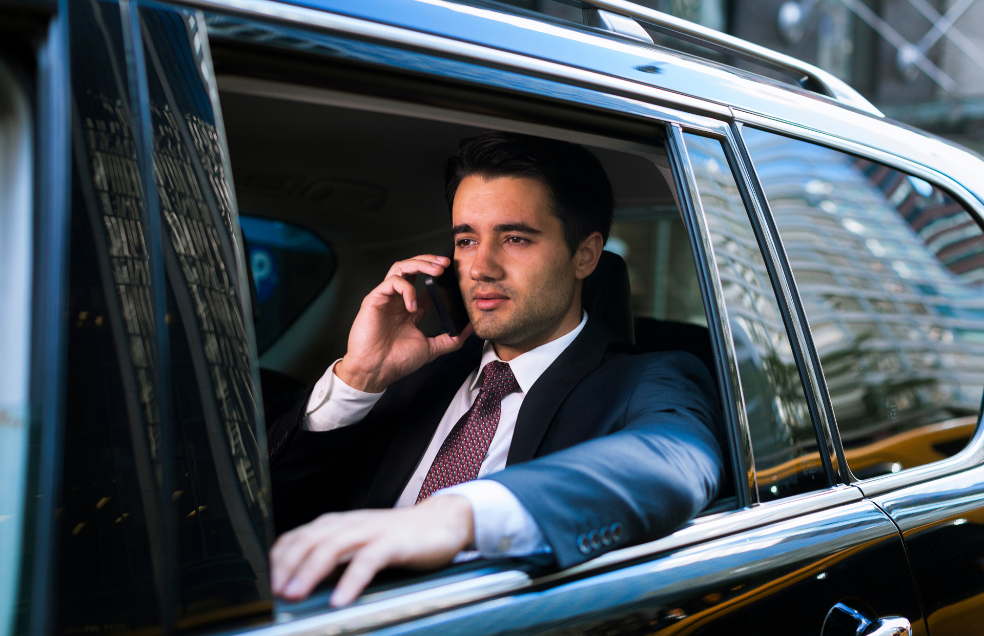 Man in a dark suit talking on a phone inside a black car, looking out the window.