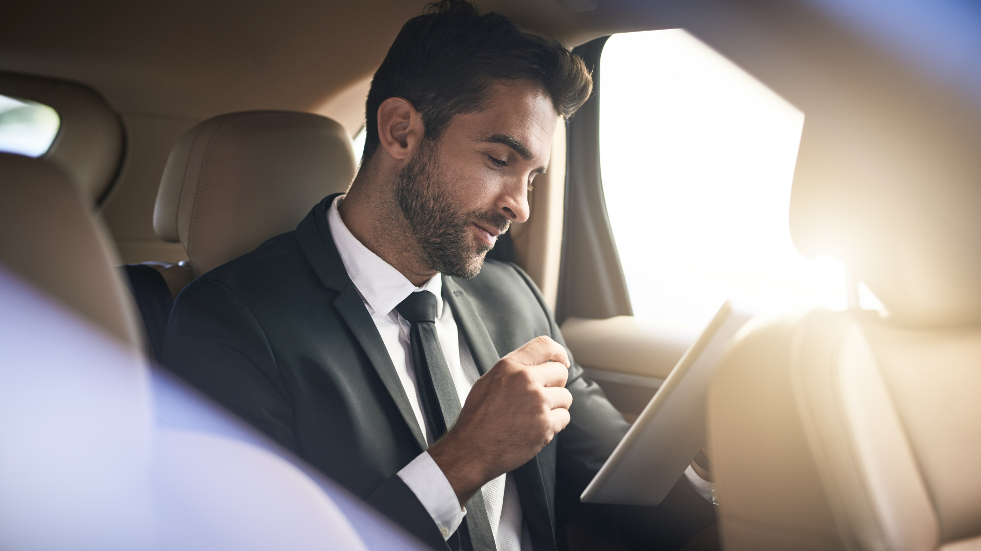 Man in suit smiling in a car, looking at a tablet in bright sunlight