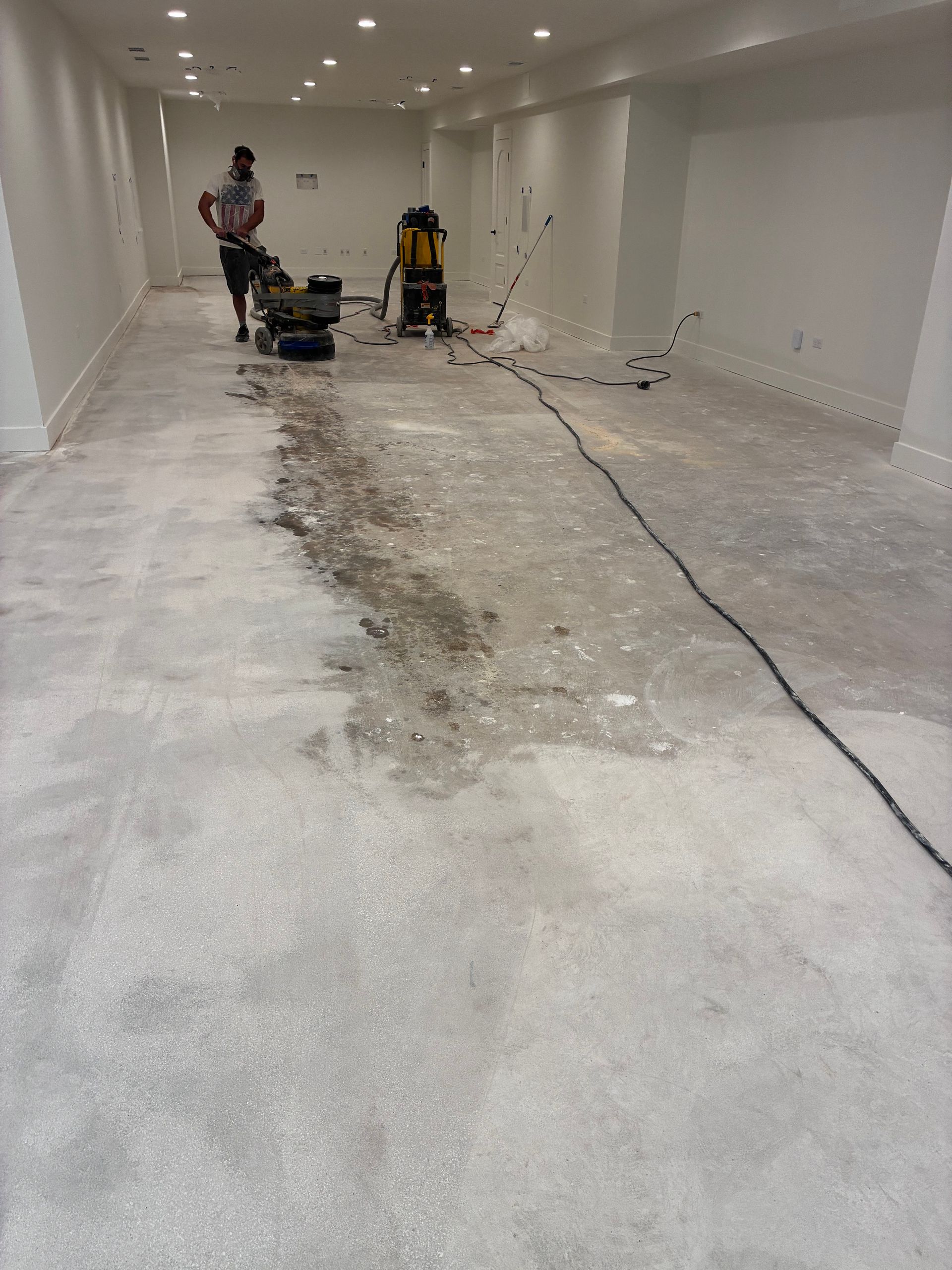 Large unfinished room with concrete floor, exposed wires, and two workers using equipment at the far end