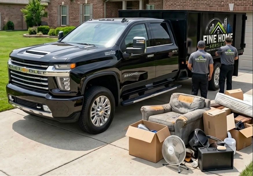 Black pickup truck parked beside a pile of household items with two people near a moving truck sign outside a home