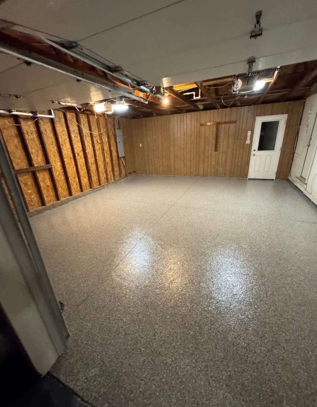 Empty unfinished basement with speckled floor, exposed ceiling, wood-paneled wall, and a white door.