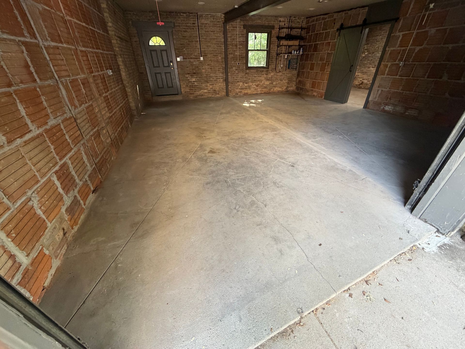 Empty unfinished room with brick walls, concrete floor, exposed wiring, and a small window and door.
