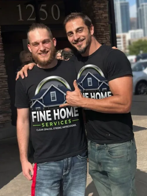 Two men in black Fine Home Services shirts smiling and posing outdoors in front of a building.