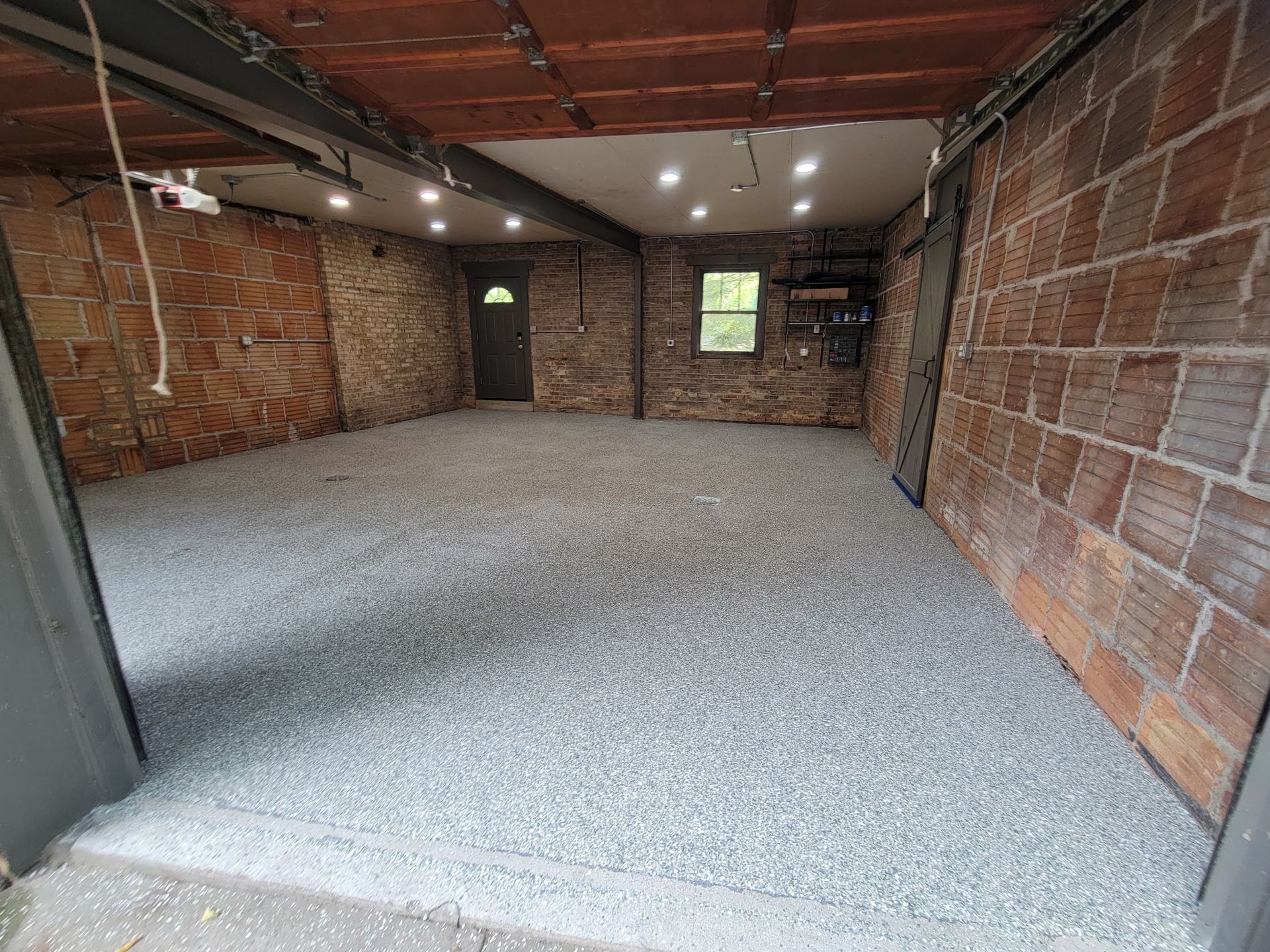 Empty unfinished room with exposed brick walls, gray carpet, and a wooden ceiling with lights.