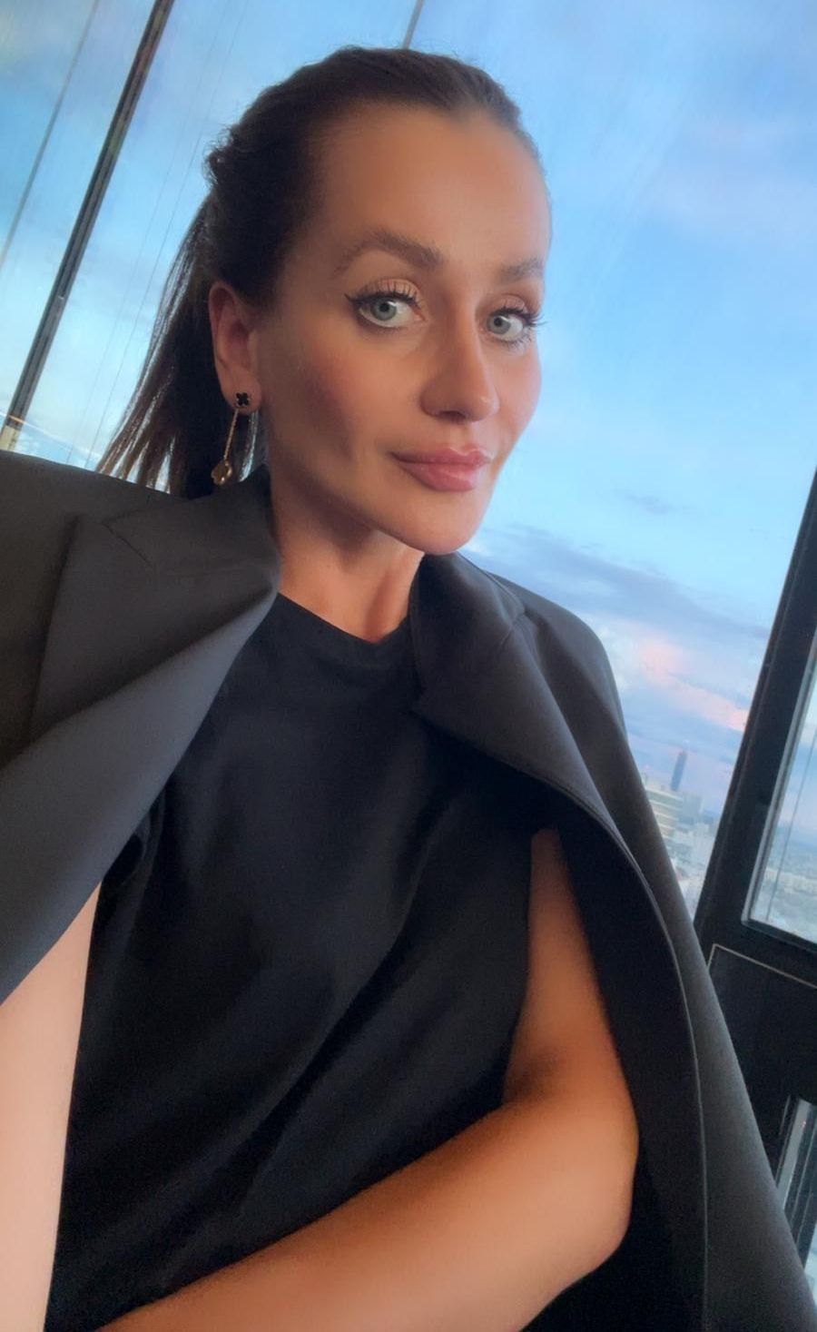 Frau in schwarzem Outfit am Fenster mit Blick auf die Stadt bei Sonnenuntergang.