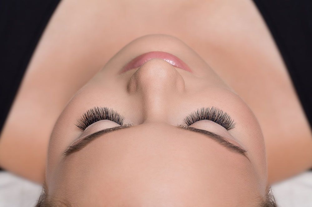 Nahaufnahme einer Person, die mit geschlossenen Augen auf dem Rücken liegt und deren Wimpern und Augenbrauen geschminkt sind.