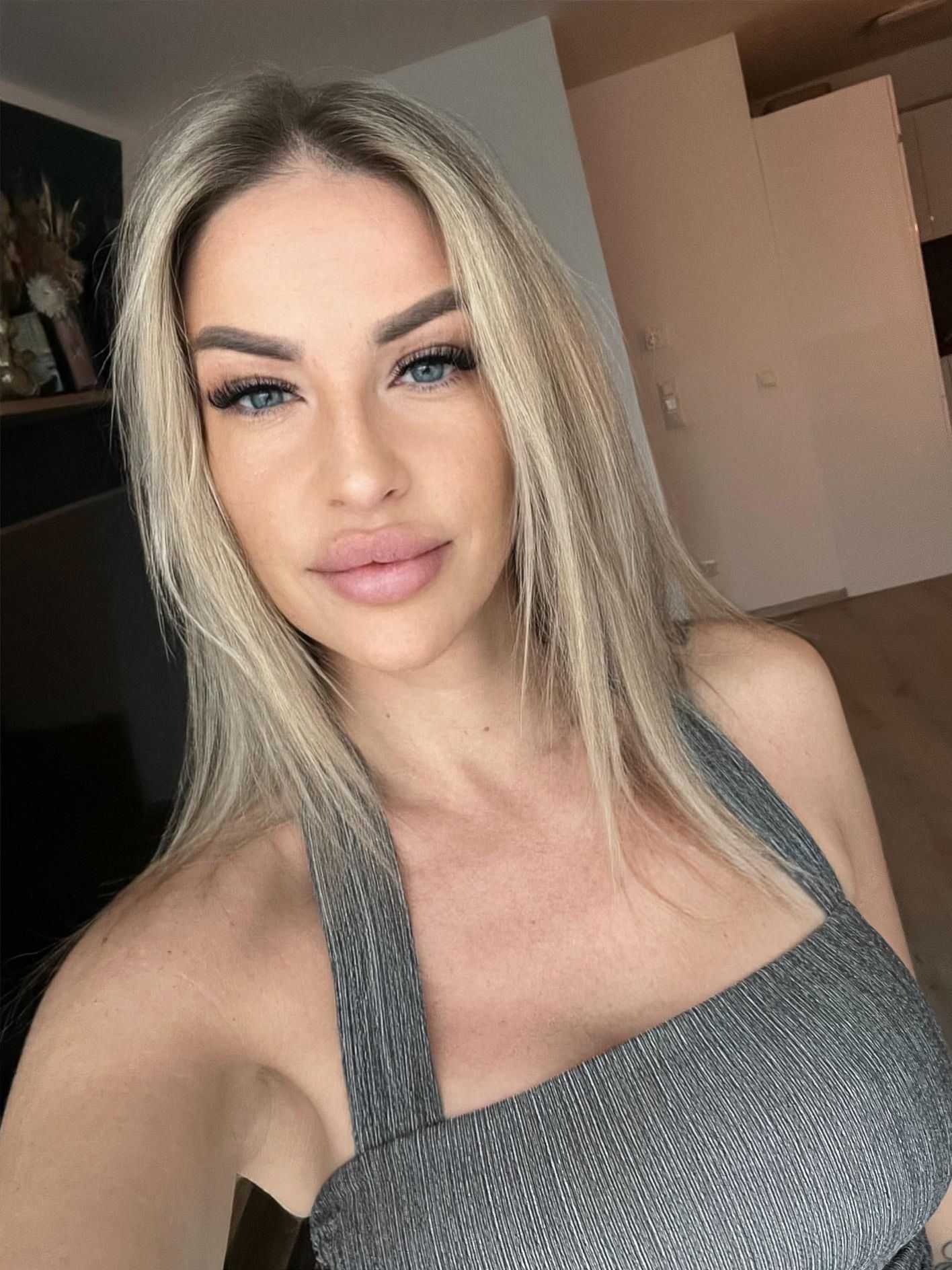 Frau in einem grauen Stricktop macht drinnen ein Selfie, mit langen blonden Haaren und neutralem Gesichtsausdruck