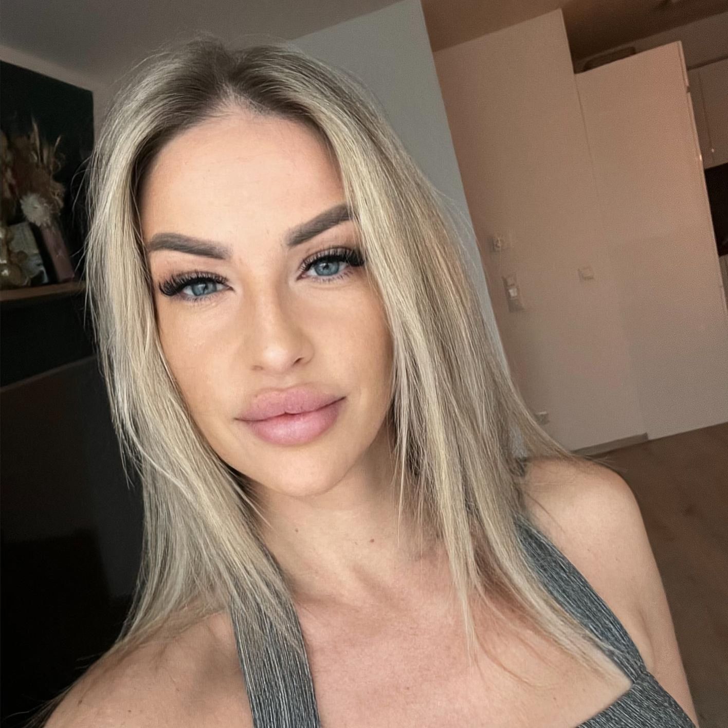 Porträt einer blonden Frau in einem grauen Oberteil, die drinnen mit neutralem Gesichtsausdruck posiert.