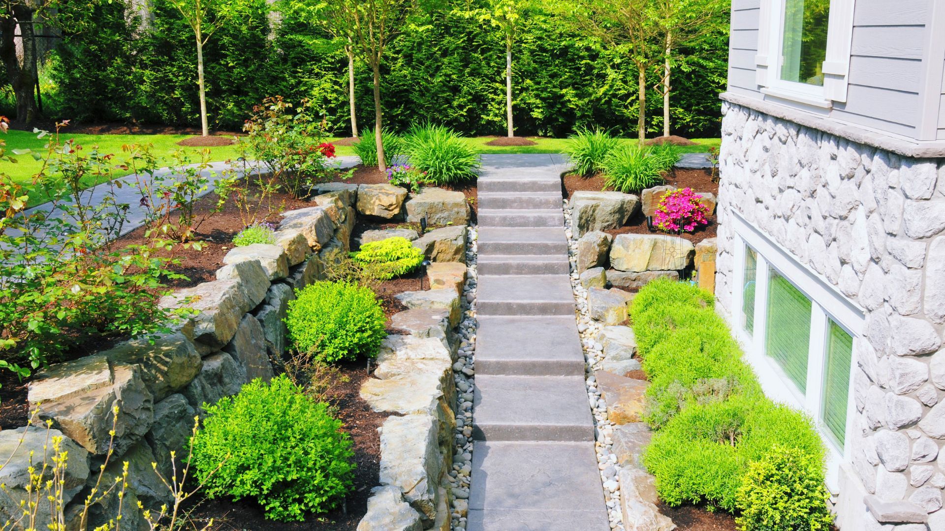 A stone staircase leads alongside a residential stone house, bordered by tiered retaining walls and green garden landscaping.