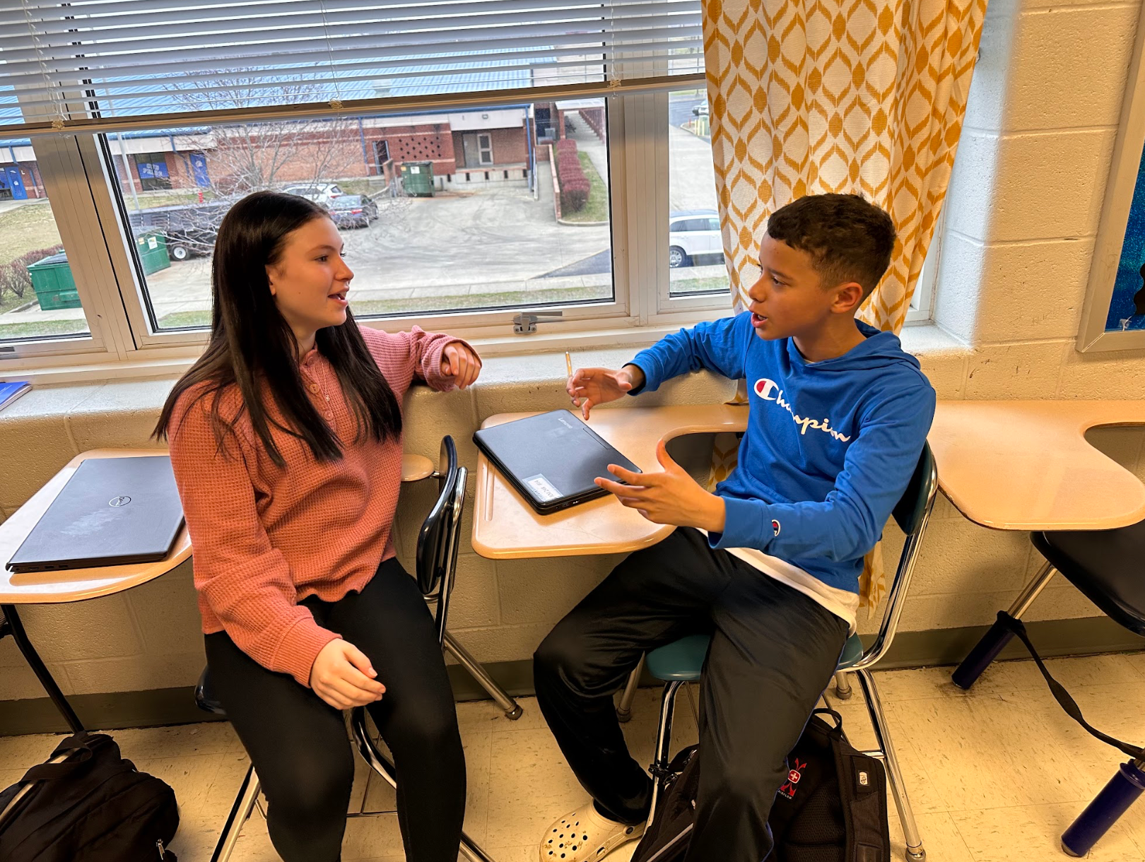 A boy and a girl are sitting at desks in a classroom talking to each other.