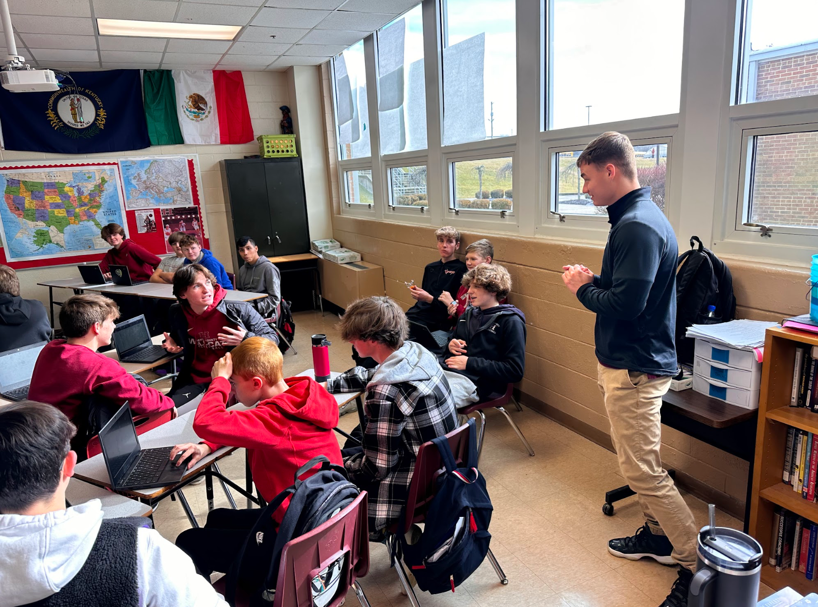 A group of students are sitting at tables in a classroom while a man stands in front of them.