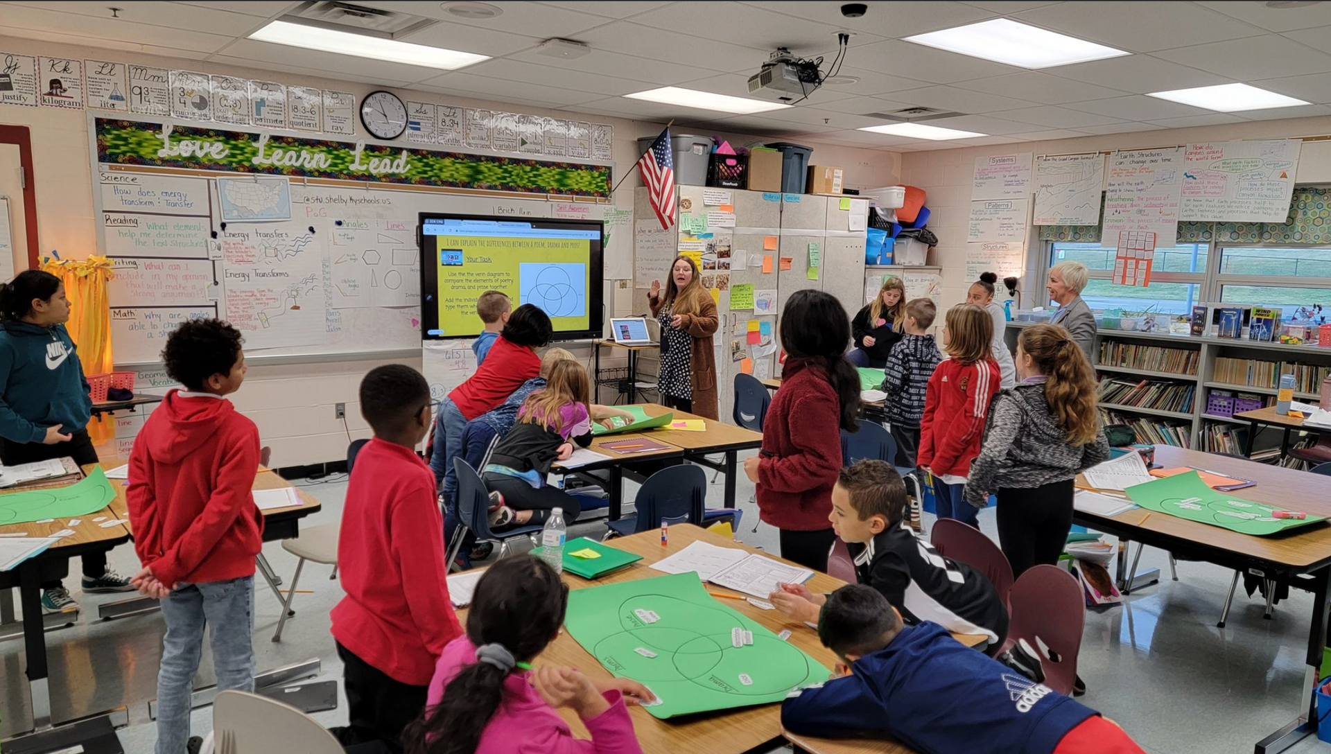 A group of children are standing around tables in a classroom.