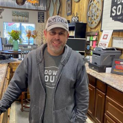 Smiling man in a gray hoodie and cap standing behind a counter in a café or shop