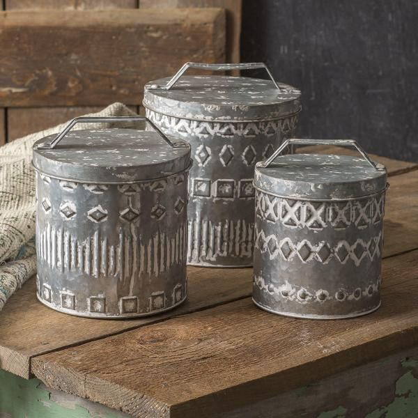 Three engraved metal canisters with lids on a wooden table, in muted gray tones.