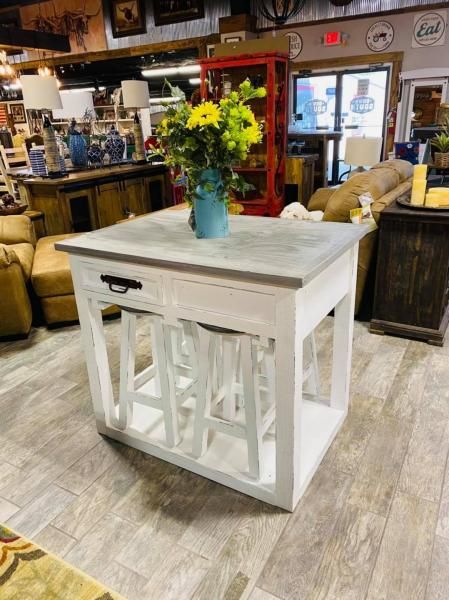 White farmhouse table with stool, topped with a blue vase of yellow flowers in a rustic store setting